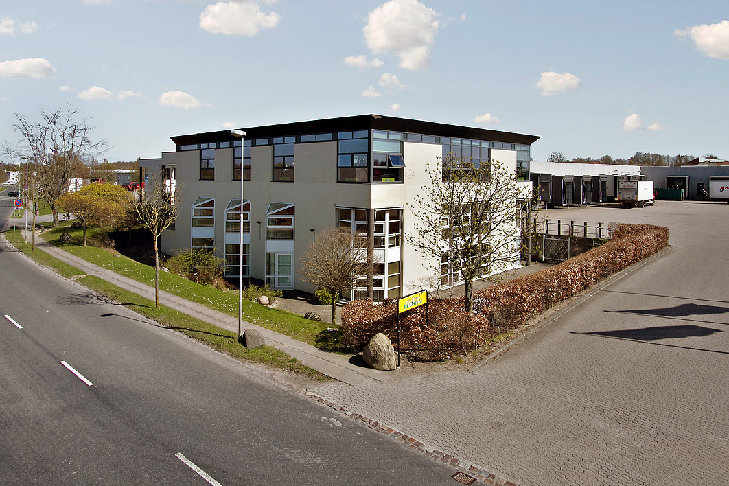 En moderne kontorbygning med store vinduer ligger på en hjørnegrund ved siden af en vej og en parkeringsplads, omgivet af træer og hække under en delvis overskyet himmel.