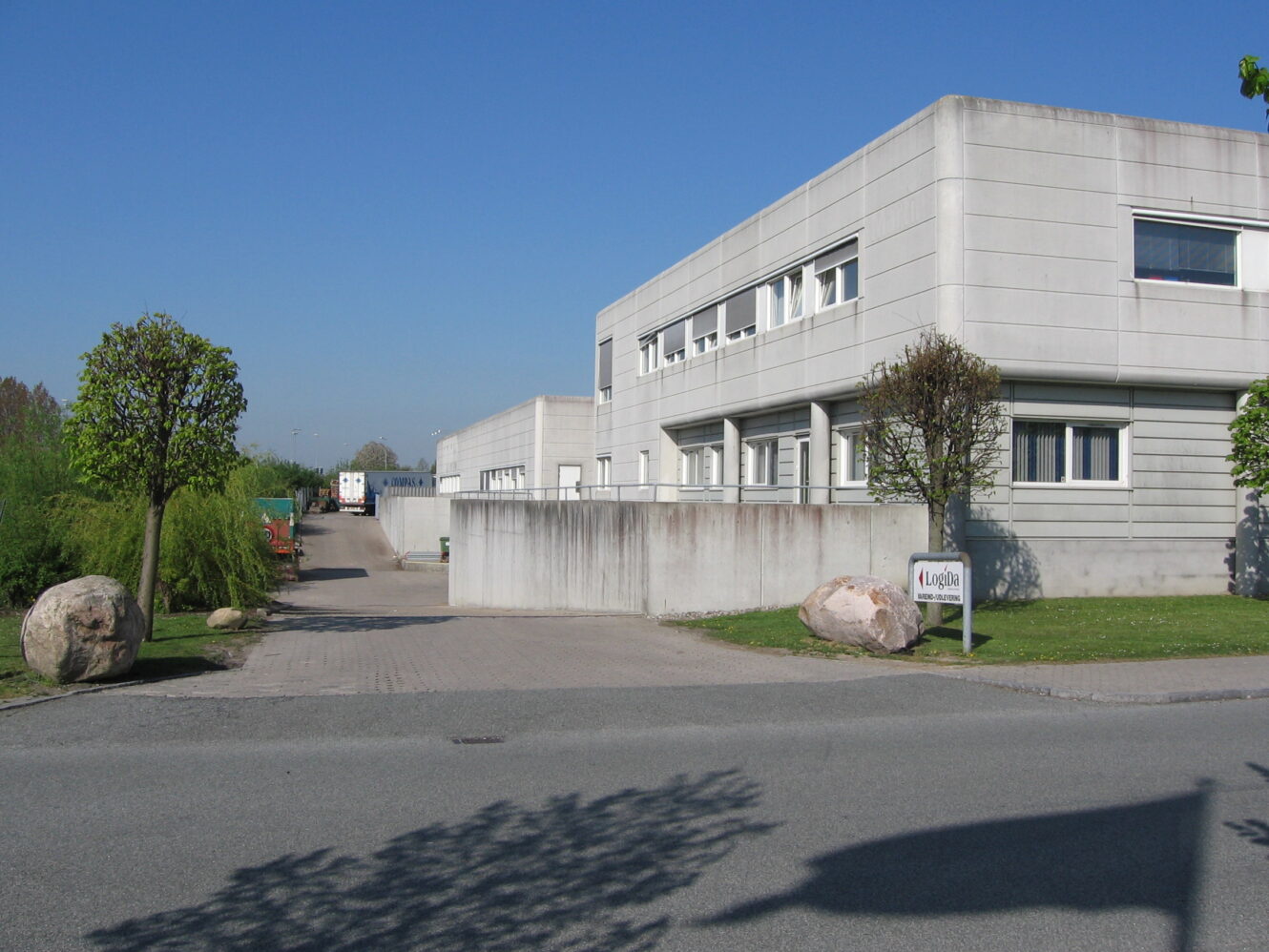Moderne industribygning med gråt ydre, omgivet af klippede træer og kampesten, under en klar blå himmel. Et skilt og en lastbil er synlige nær indgangen.