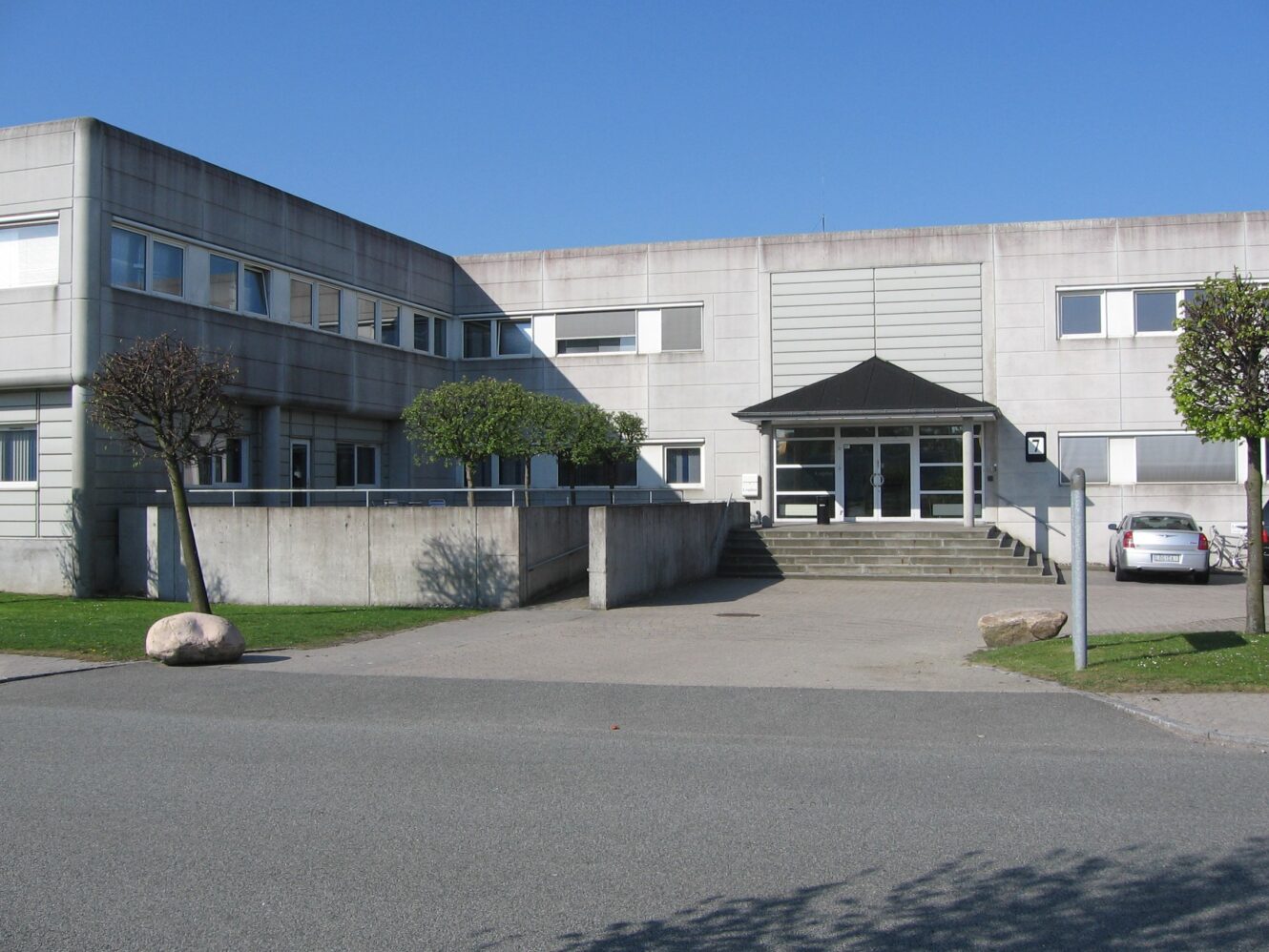 En moderne, toetagers grå bygning med fladt tag, store vinduer og en trappeformet indgang under en lille baldakin, fotograferet på en klar dag.