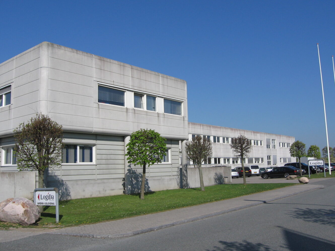 En moderne, grå kontorbygning med rektangulære vinduer, små træer og parkerede biler udenfor. På skiltene nær indgangen står der Logica. Klar blå himmel over hovedet.