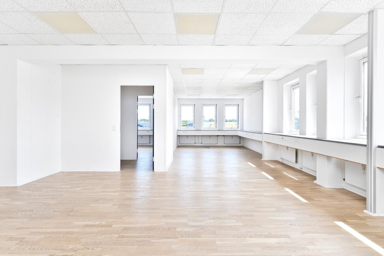 Lyst, tomt kontorlokale med store vinduer, hvide vægge og lyse trægulve. Vinduer langs højre side lader naturligt lys fylde rummet.