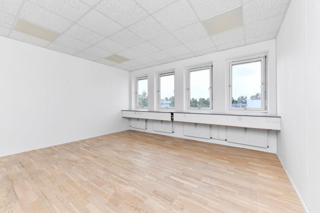 Et tomt rum med hvide vægge, lyse trægulve, nedhængt loft og fire store vinduer langs den ene væg, der lukker naturligt lys ind.