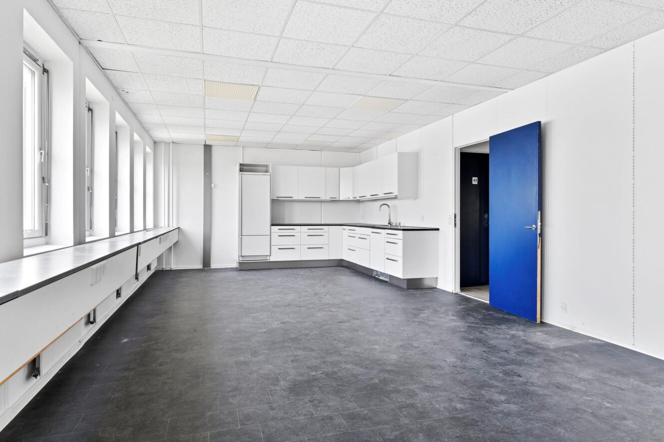 Stort, tomt rum med en række vinduer, hvide vægge, sort gulv, et tekøkken langs den ene væg og en blå, åben dør.