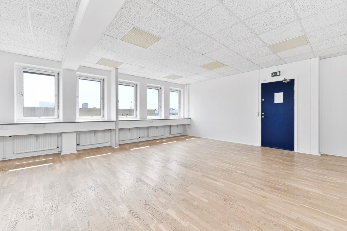 Tomt, lyst rum med lyst trægulv, hvide vægge, fire store vinduer, nedhængte loftspaneler og en blå dør.