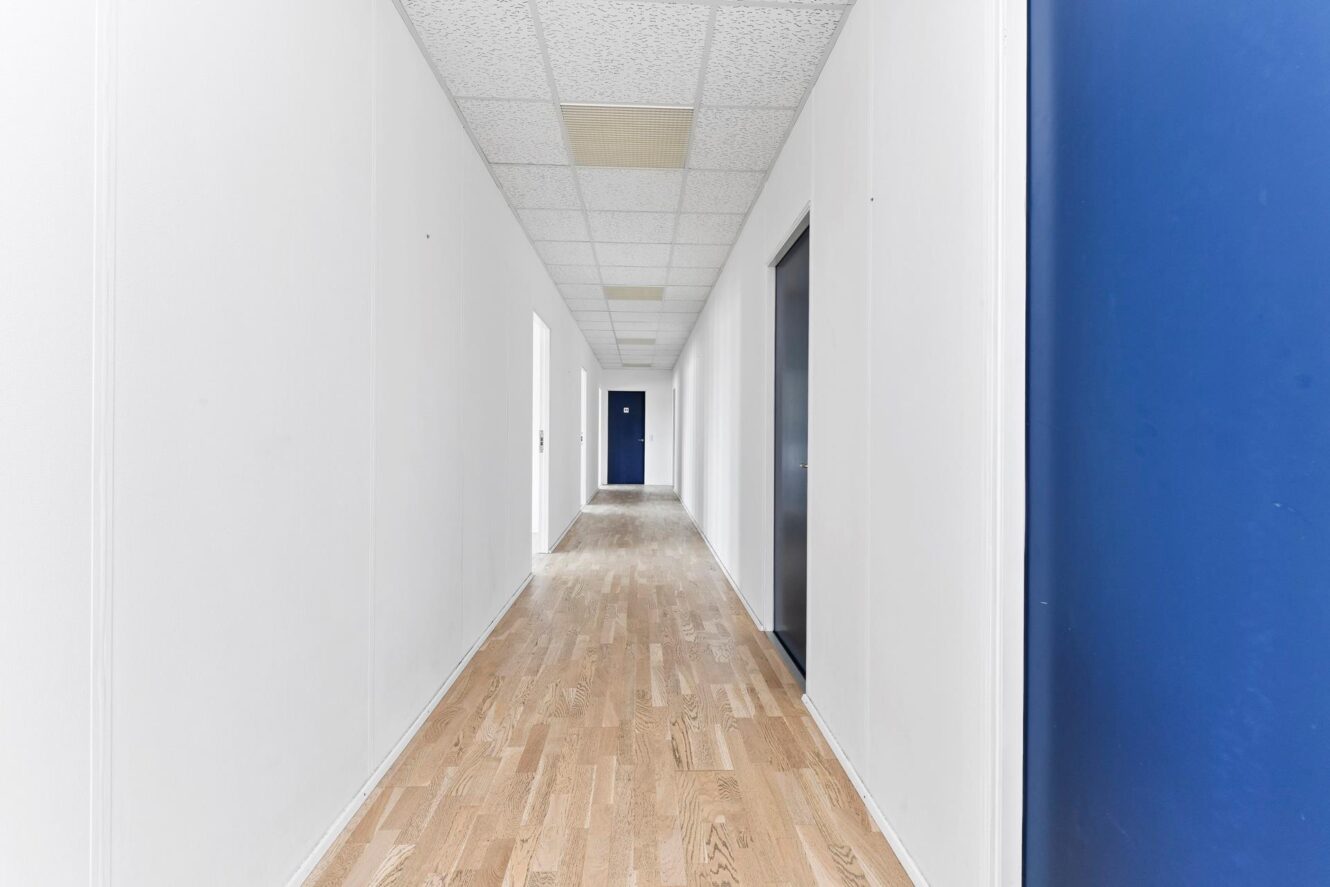 En lang, tom gang med hvide vægge, et lyst trægulv og døre med blå rammer i højre side. Loftpladerne er synlige.