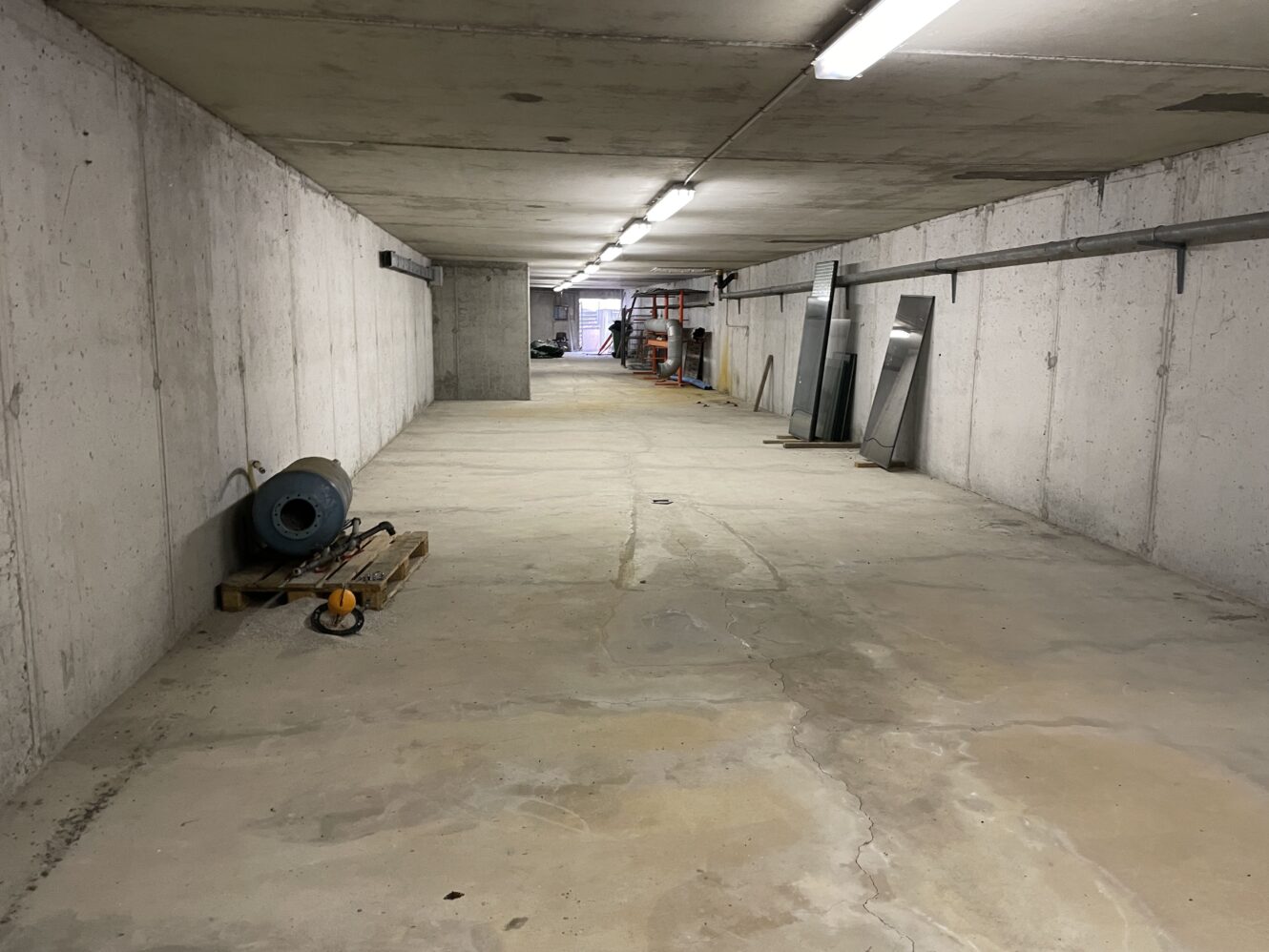 Lang, tom betonkælder med blottede vægge, loftslamper, spredte byggematerialer og hylder i den fjerneste ende.