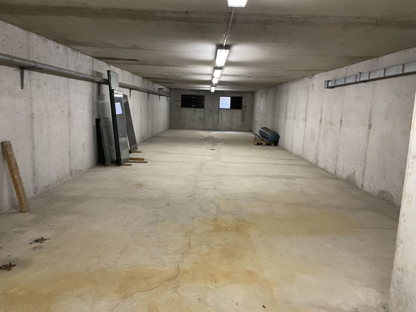 Tom betonkælder eller garage med lysstofrør over hovedet, et par ting mod venstre væg og små vinduer i den fjerneste ende.
