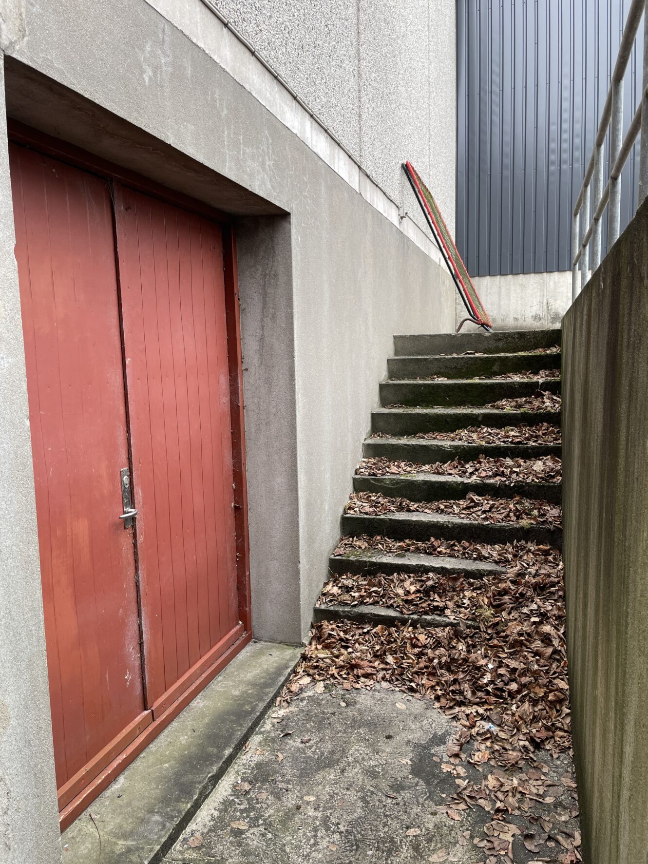 Udendørs betontrapper dækket af tørre blade fører op ved siden af en rød dør i en grå betonbygning; en rød og hvid pæl hviler på trappens gelænder.