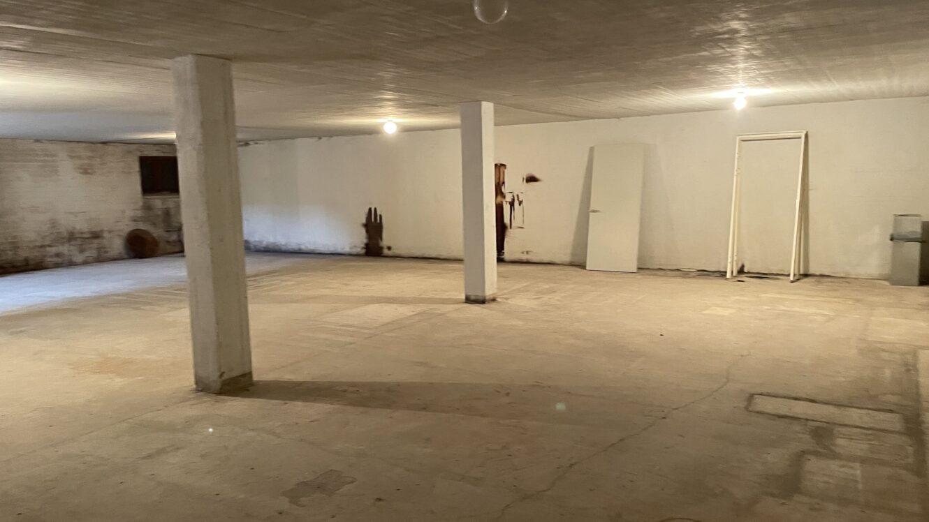 Stor tom kælder med betongulv, hvide vægge, synlige søjler, et lille vindue, nogle pletter på væggen, en dør, der læner sig op ad væggen, og synlige rør i det ene hjørne.