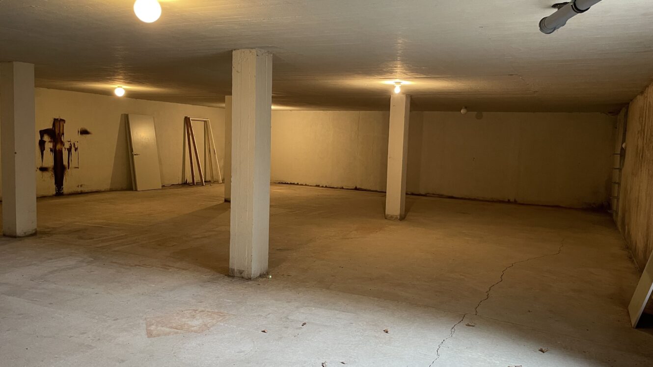 Stor tom kælder med bart betongulv, støttesøjler, blottede lyspærer og et par døre og brædder, der læner sig op ad væggen.