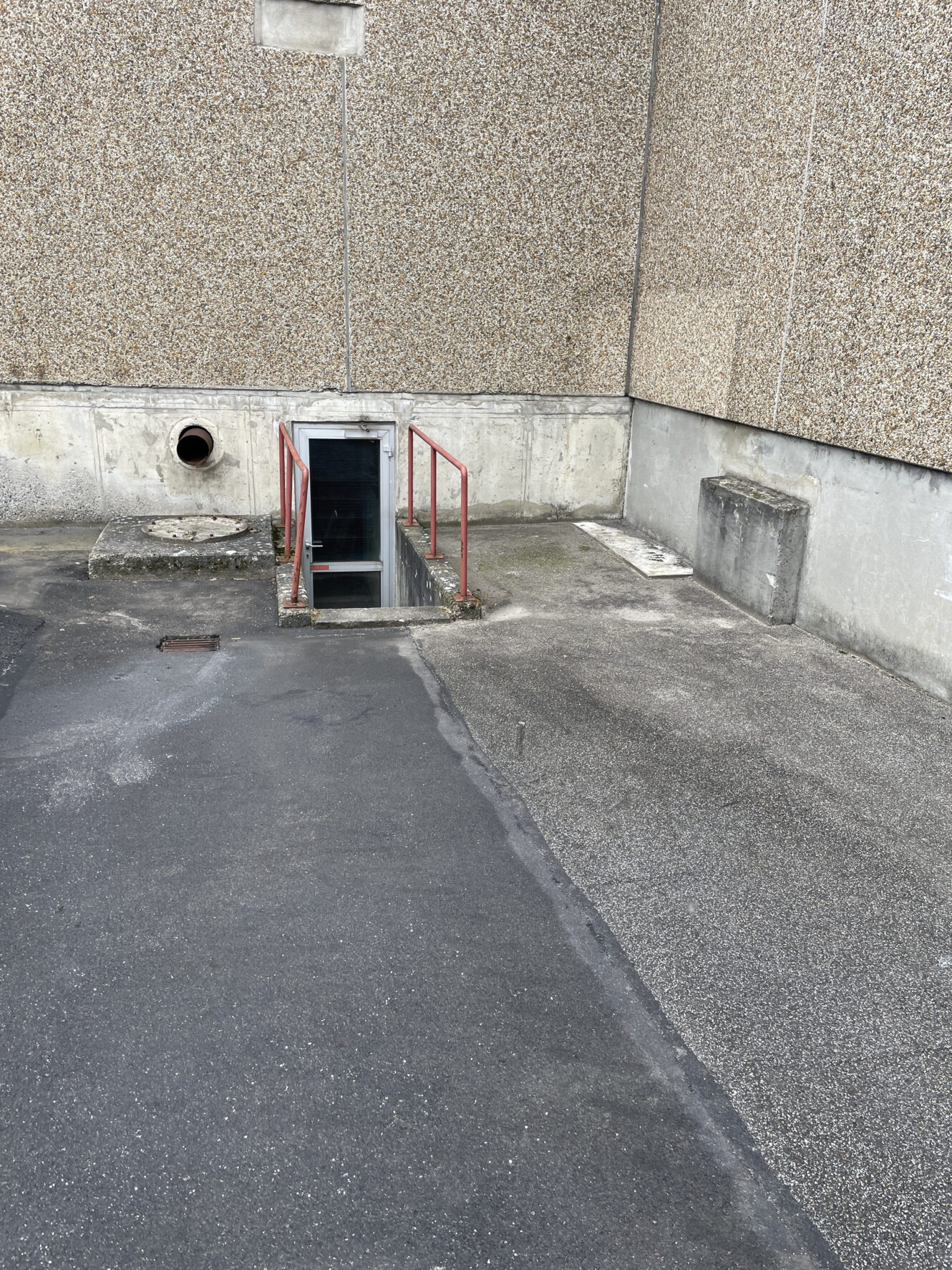 Et udendørs betonområde med en trappe, der fører ned under jorden, omkranset af røde rækværk, der støder op til en plettet bygningsmur.