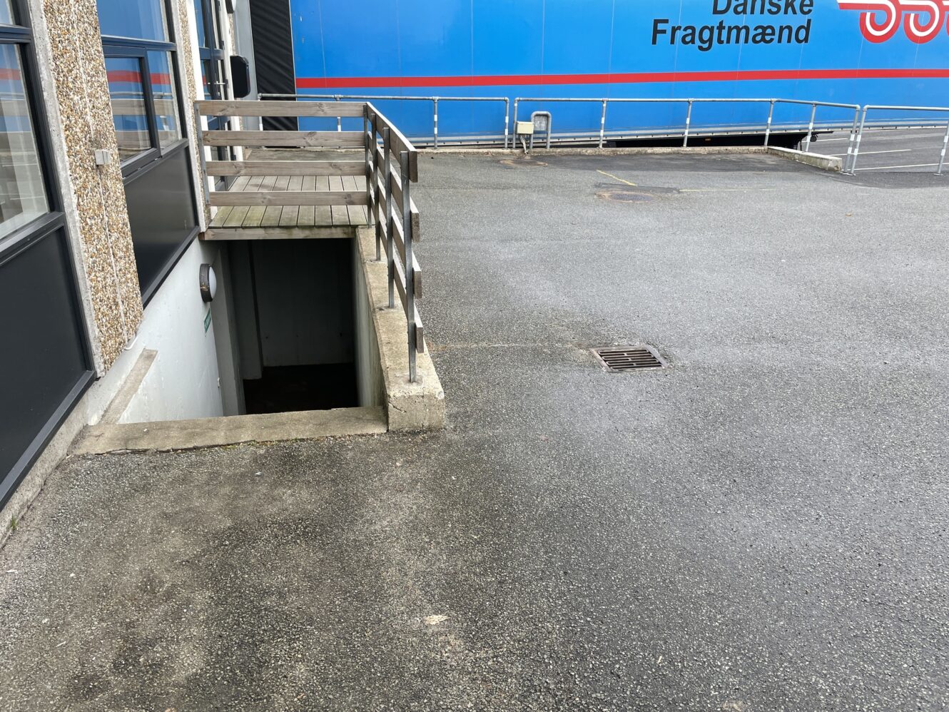 En trappe fører ned fra en betonbro ved siden af en bygning. En blå lastbil med Danske Fragtmænd holder parkeret i baggrunden.