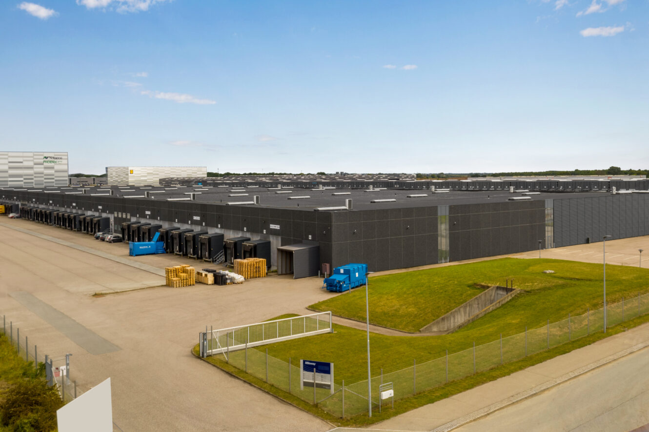 Stort industrielt lagerkompleks med flere læsseramper, lastbiler, stablede paller og et indhegnet græsareal under en klar himmel.