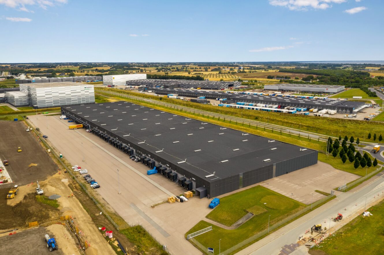 Luftfoto af et stort industrilager med flere læsseramper, omgivet af parkeringspladser, veje og grønne områder på en klar dag.