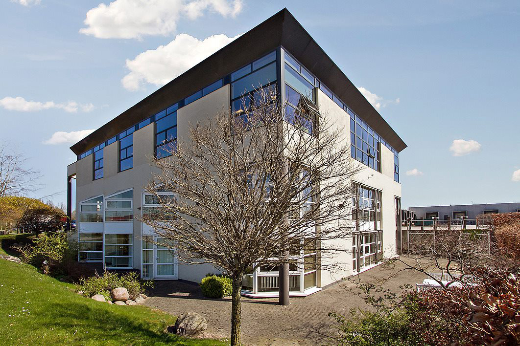 Moderne kontorbygning i tre etager med store vinduer og et trekantet tag, omgivet af en asfalteret sti, græs og bladløse træer under en blå himmel med spredte skyer.