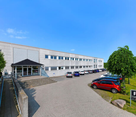 En moderne erhvervsbygning i to etager med store vinduer, en hovedindgang med trapper og en parkeringsplads med flere biler under en klar blå himmel.