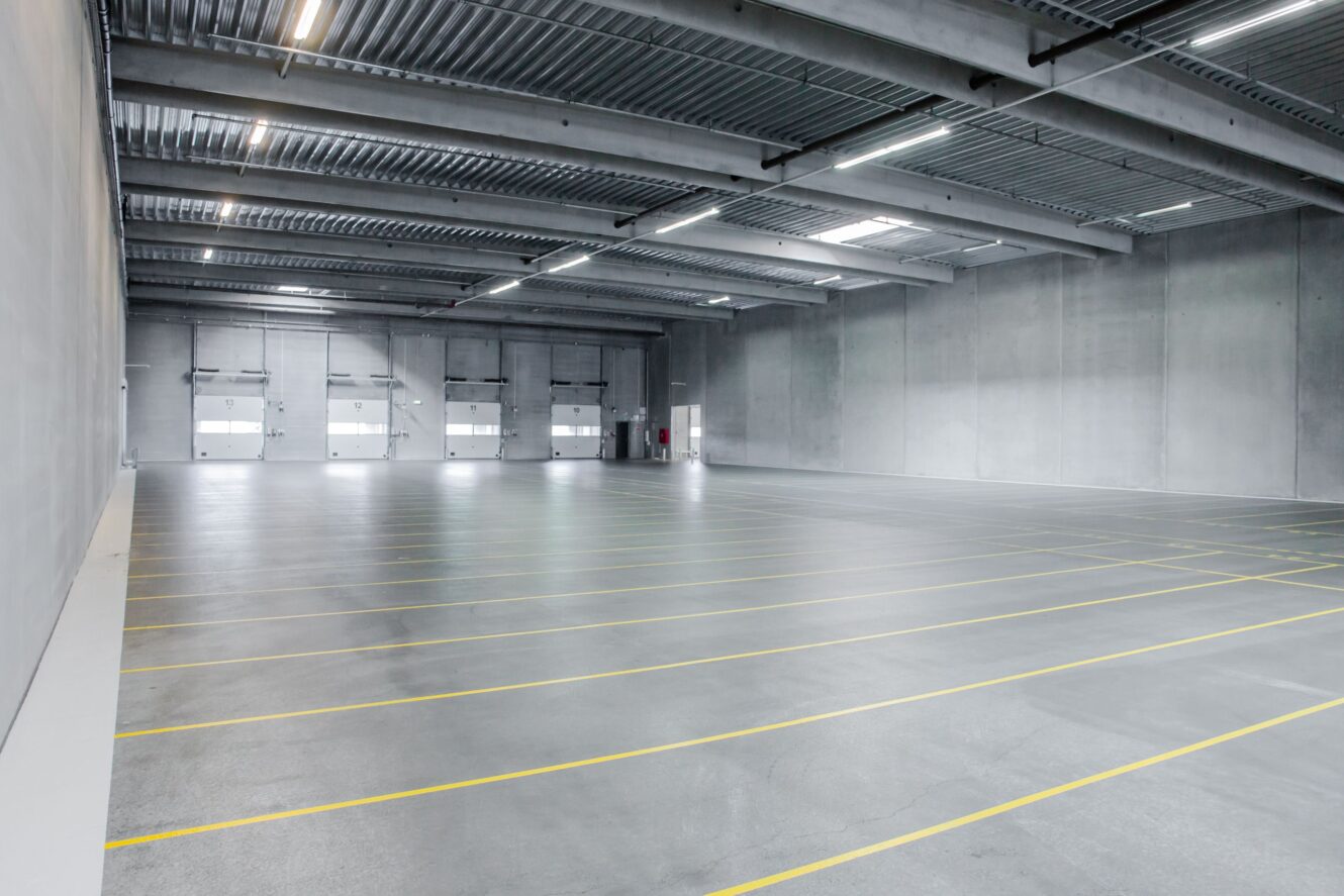Interiør af et tomt industrilager med højt til loftet, betongulve, grå vægge, ovenlys og markerede gule linjer til parkering eller lagerplads.