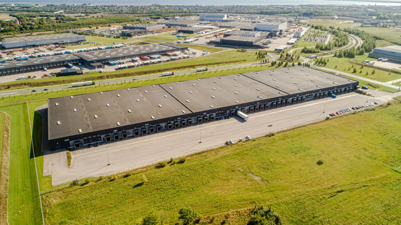 Luftfoto af et stort industrilager med flere læsseramper, omgivet af marker og andre erhvervsbygninger.