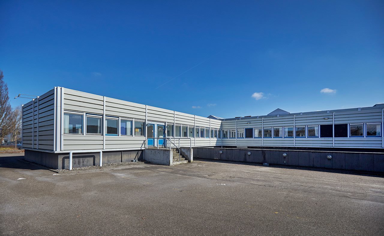 Industribygning i én etage med fladt tag, store vinduer og en blå indgangsdør under en klar blå himmel med et tomt asfalteret område foran.