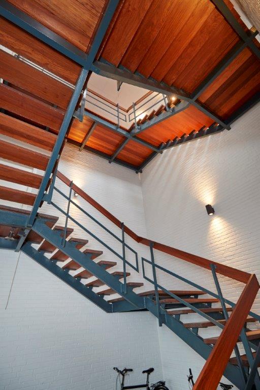 Indendørs trappe med metalramme og trætrin, vinklet opad i flere trin, mod hvide murstensvægge med vægmonterede lamper. To cykler er synlige i bunden.