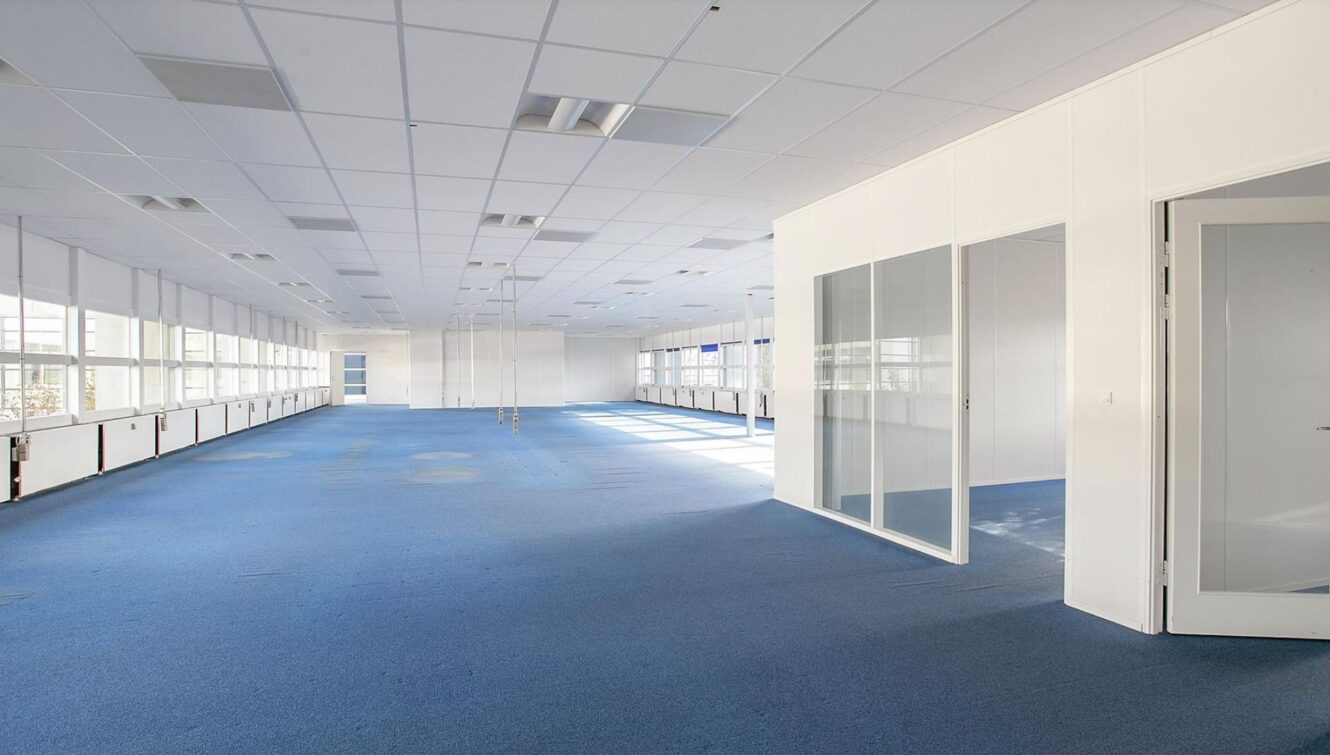 Rummeligt, tomt kontor med blåt gulvtæppe, hvide vægge, glasvægge og store vinduer, der lukker naturligt lys ind.