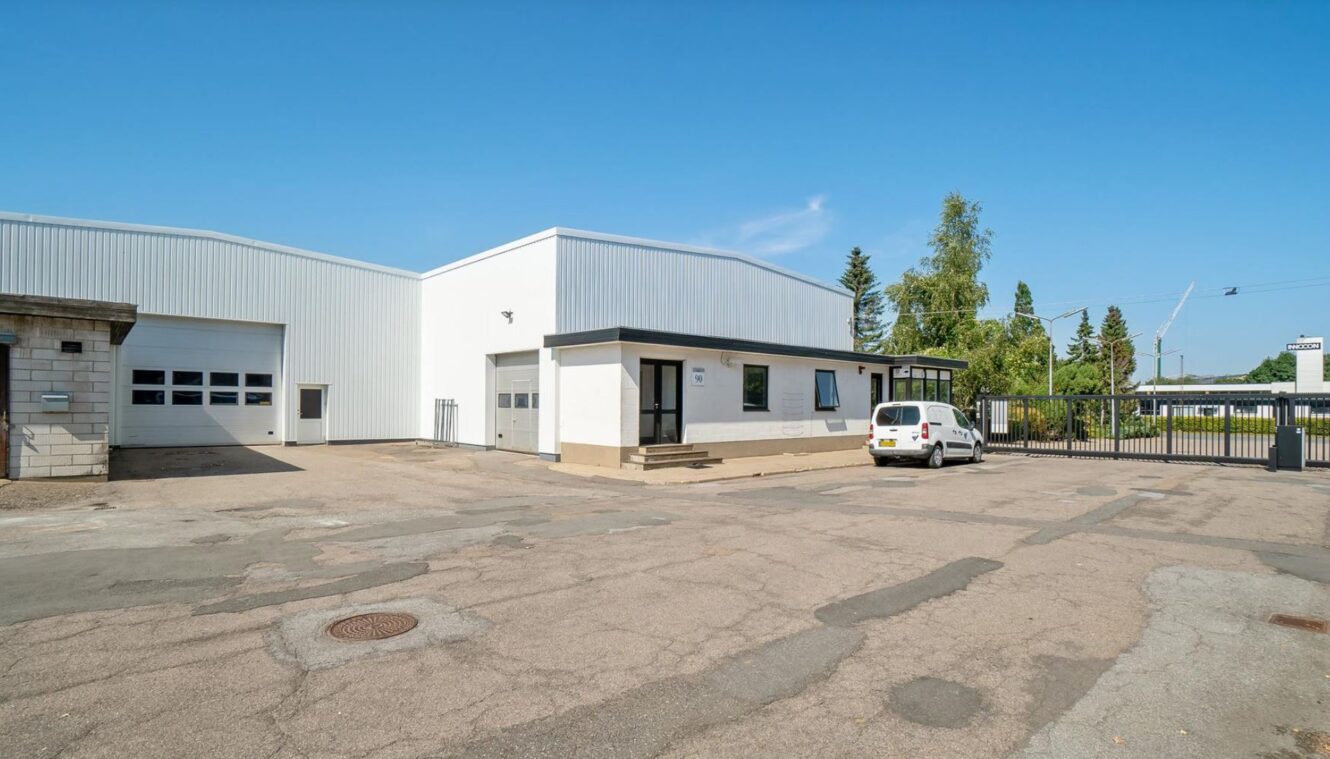 En hvid industriel lagerbygning med en lille kontorindgang, parkeringsplads, en parkeret hvid varevogn og et indhegnet område på en klar, solrig dag.