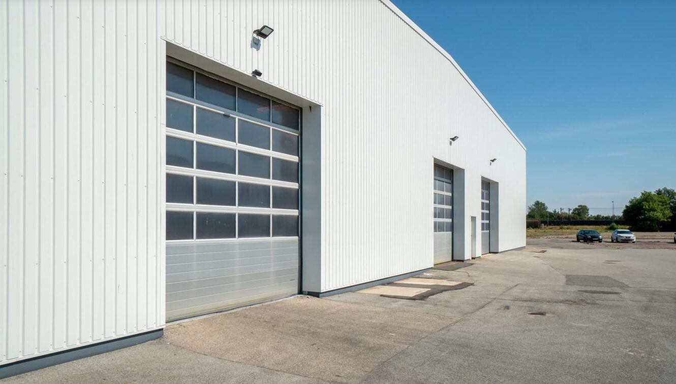 Stort hvidt industrilager med tre lukkede garageporte og udendørs belysning, beliggende på en asfalteret grund under en klar blå himmel. Et par træer og en bil er synlige i baggrunden.