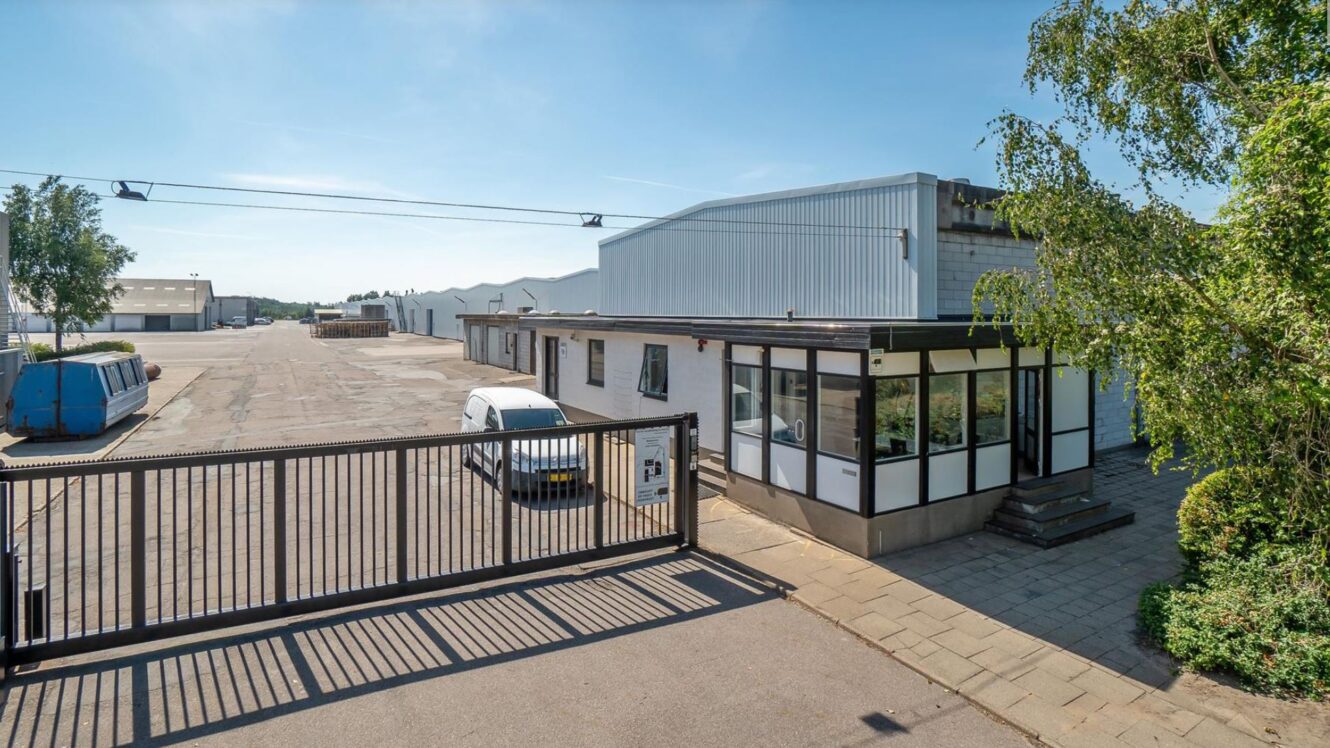 Et indhegnet industrianlæg med en lille kontorbygning, en parkeret hvid bil, affaldscontainere og store lagerbygninger i baggrunden på en klar dag.