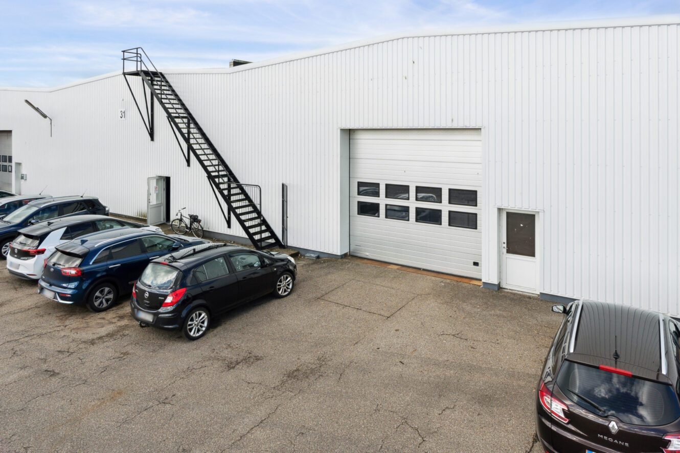 Udsigt til en hvid industribygning med en stor garageport, en lille sidedør, en udvendig metaltrappe og flere parkerede biler.