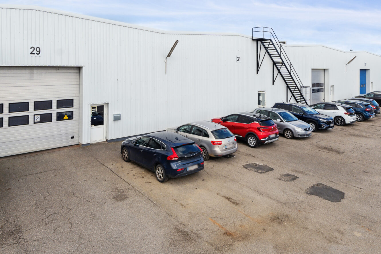 Flere biler er parkeret foran en hvid industribygning med nummererede garageporte og en udvendig metaltrappe.