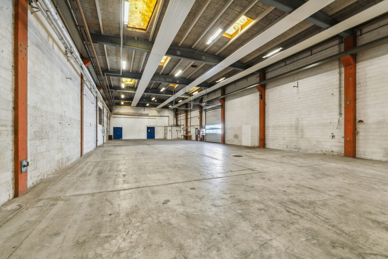 Bredt billede af et tomt industrilager med betongulve, højt til loftet, synlige bjælker og hvide murstensvægge; fluorescerende lys og blå døre er synlige.