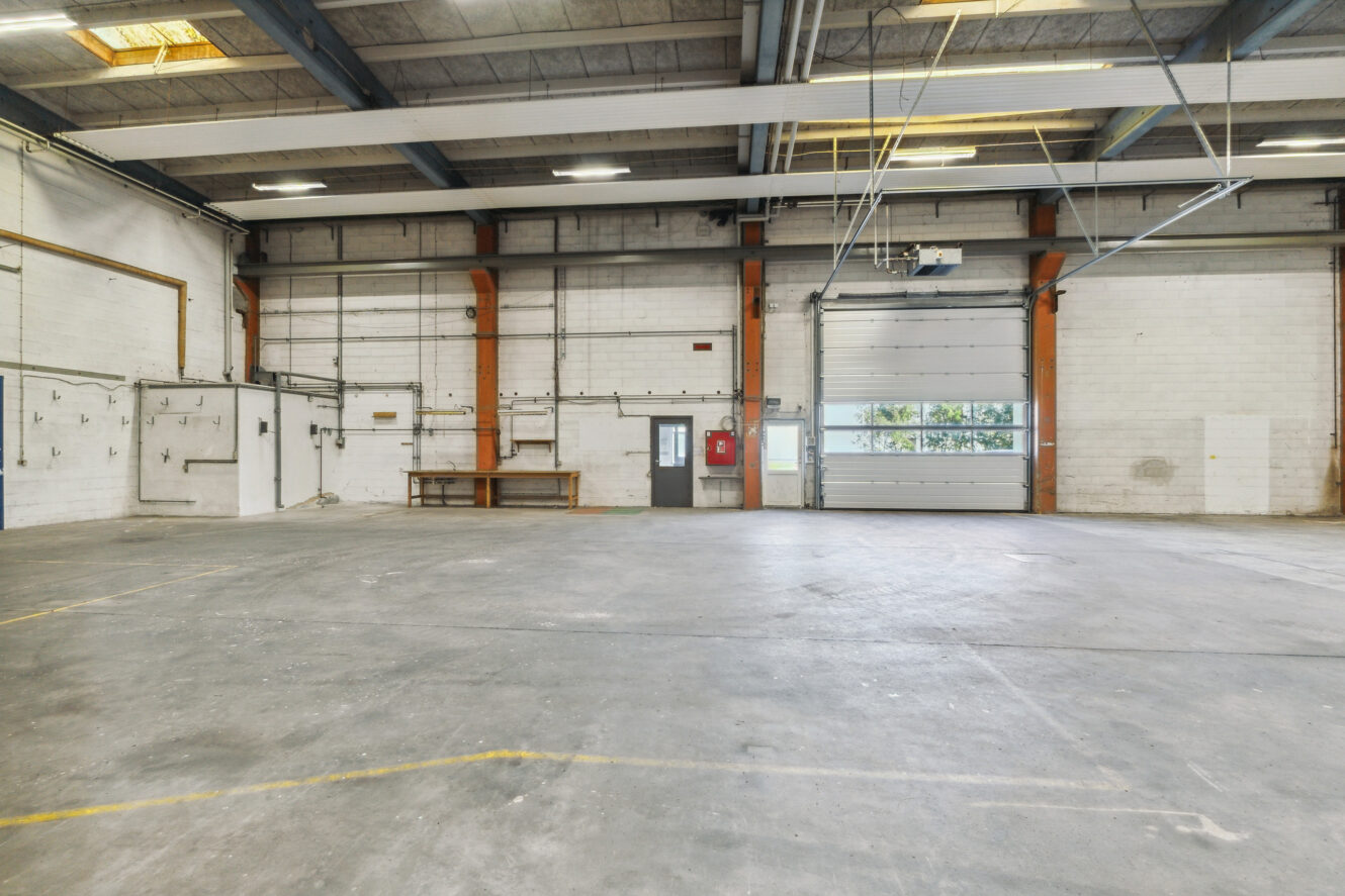 Rummelig tom lagerhal med betongulv, højt til loftet, hvide murstensvægge, stor garageport, arbejdsbænk og ovenlys.