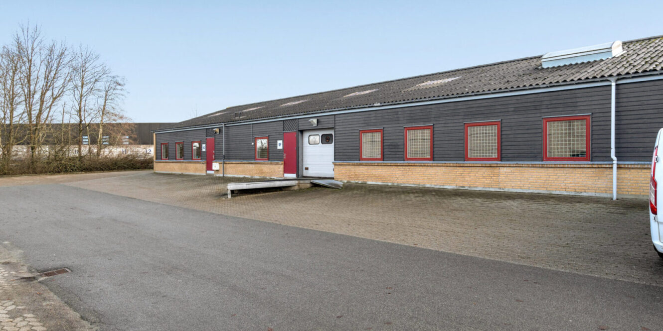 Industribygning i én etage med mørk facadebeklædning, røde vinduer, en central dør med rampe og et asfalteret område foran. Træer er synlige til venstre.