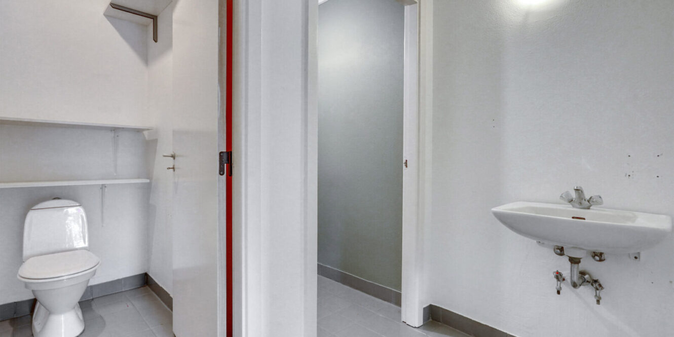 Et toilet med et toilet i en separat bås, åbne hylder, en væghængt vask og synlige rør under vasken.