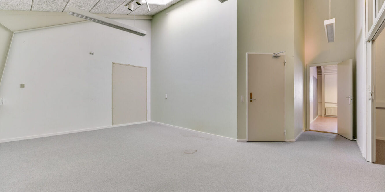 Tomt hvidt rum med lysegråt gulvtæppe, højt til loftet, lukkede og åbne døre og en gang, der er synlig gennem den åbne døråbning.