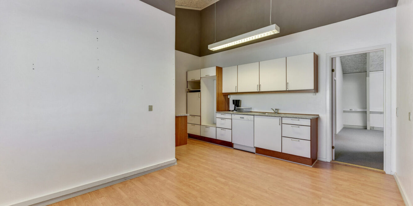 Et lille, umøbleret køkken med hvide skabe, opvaskemaskine, kaffemaskine, lyst trægulv og en tilstødende døråbning, der fører ind til et rum med gulvtæppe.