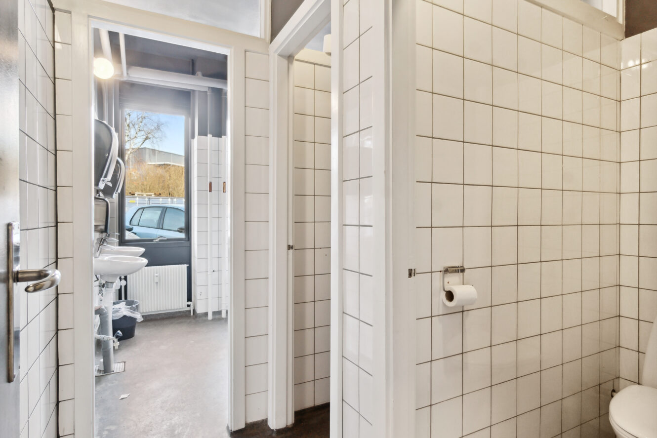 Et flisebadeværelse med separate båse, et toilet med en rulle papir og en åben dør, der afslører en vask, et spejl og et vindue med udsigt til en parkeret bil udenfor.