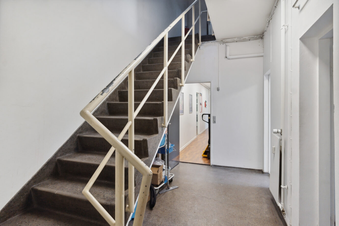 En betontrappe med et hvidt metalgelænder ved siden af en gang i en industri- eller institutionsbygning; rengøringsartikler opbevares under trappen.