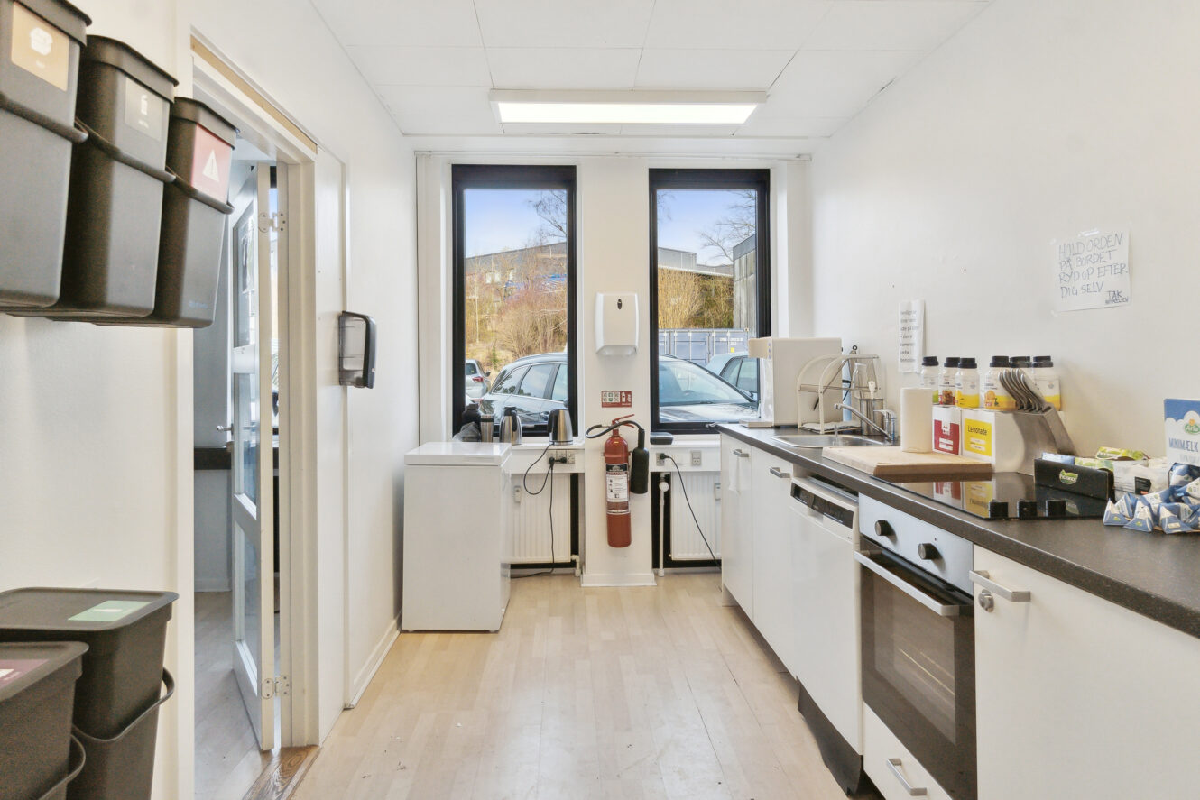 Et rent tekøkken med opvaskemaskine, vask, drikkevaredispensere og opbevaringsbeholdere. Store vinduer og en brandslukker på den fjerneste væg.