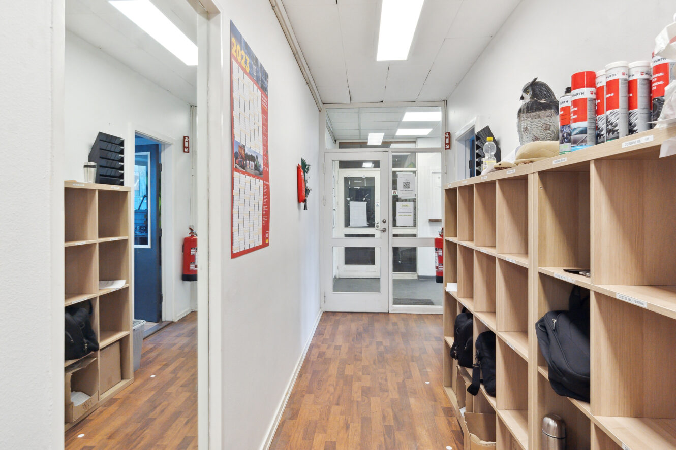 En gang med træhylder til højre, en vægkalender og en brandslukker til venstre og glasdøre for enden.