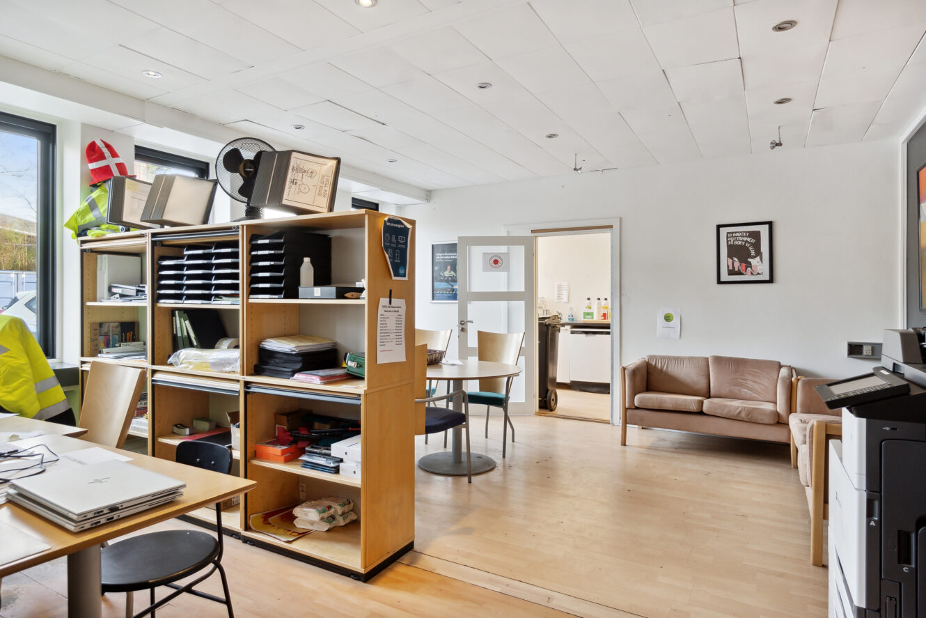 Et kontorlokale med trægulv, hylder med kontorartikler, skriveborde, en kopimaskine, en beige sofa og et tekøkken i baggrunden.