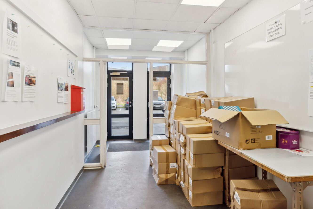 En gang med stablede papkasser til højre, et bord med kontorartikler og en glasdør, der fører udenfor for enden.