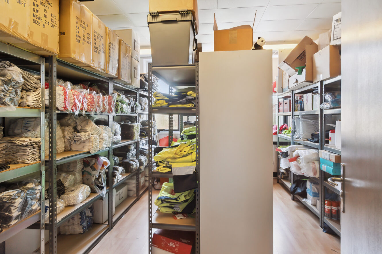 Et opbevaringsrum med metalhylder med forskellige kasser, tasker og sammenfoldet tøj, organiseret langs begge vægge og nogle stablet helt op til loftet.