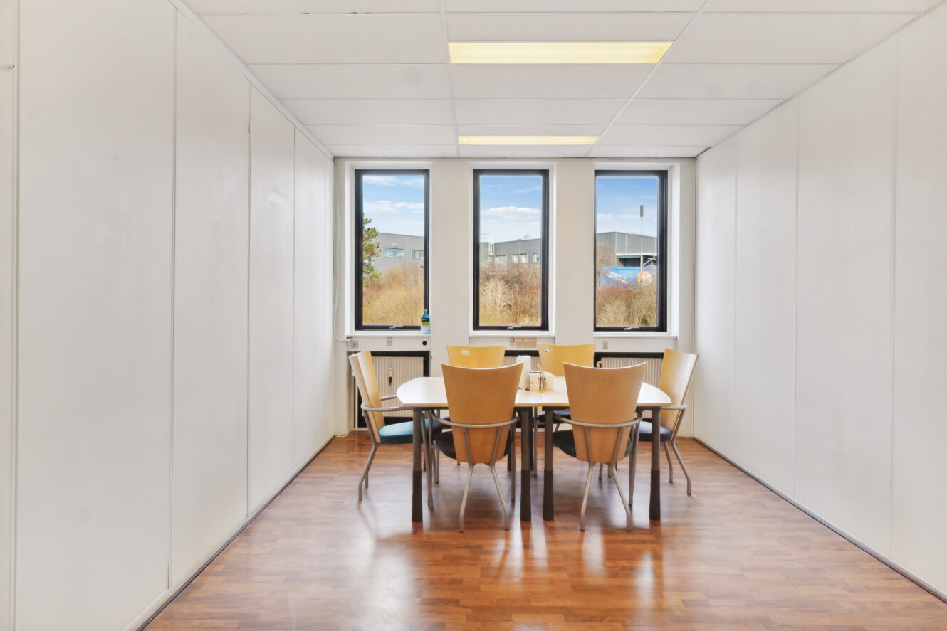 Lille mødelokale med træbord og seks stole, tre vinduer på den fjerneste væg og hvide panelvægge med trægulv.