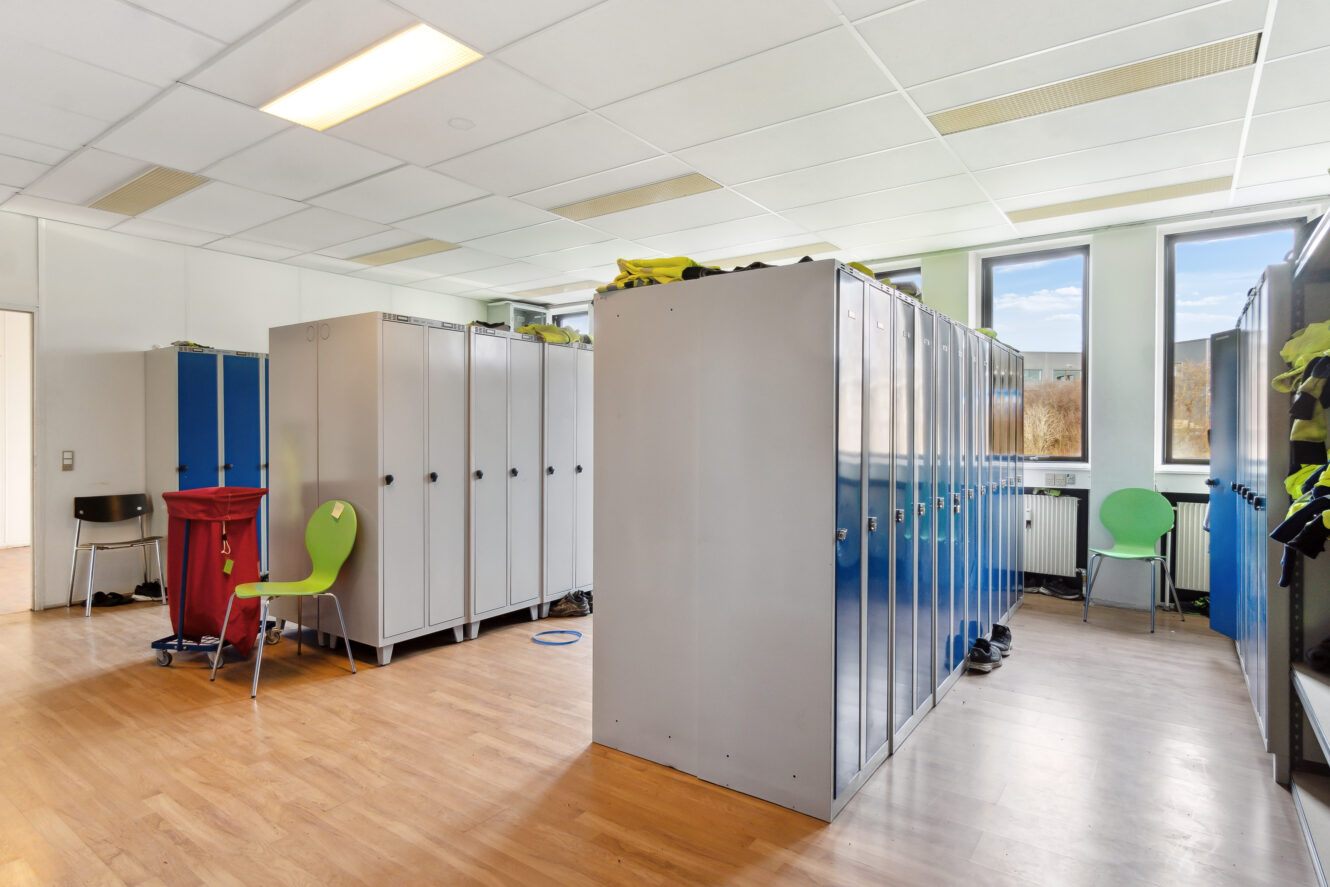 Et omklædningsrum med blå og grå skabe, grønne stole, en rød vasketøjsspand og vinduer, der lukker naturligt lys ind på et trægulv.