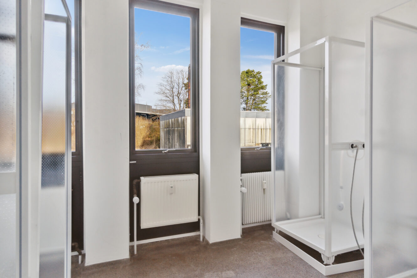 Et lyst badeværelse med to brusekabiner, store vinduer, hvide vægge og radiatorer under vinduerne.