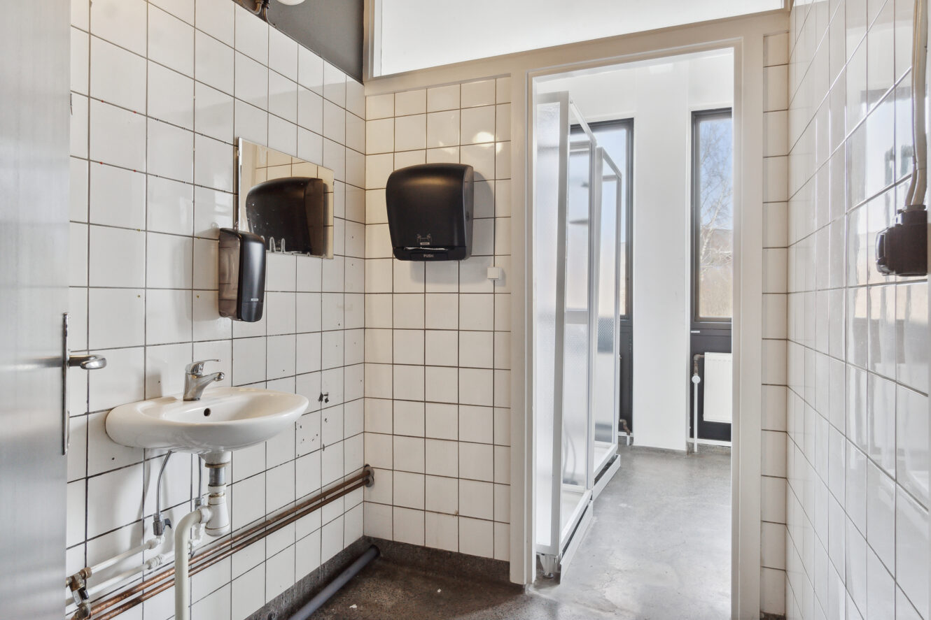 Et offentligt toilet med hvide flisebelagte vægge, en hvid håndvask, papirhåndklædedispensere og en åben dør, der fører til et bruseområde med glasskillevægge.