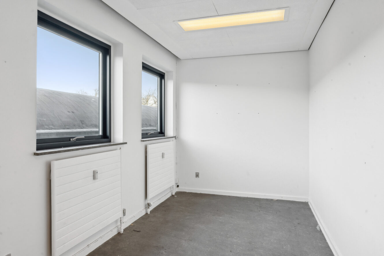 Lille, tomt hvidt rum med to vinduer, to radiatorer under dem, et gråt gulv og en rektangulær loftslampe.
