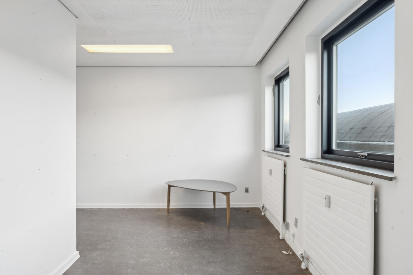 Lille, tomt rum med hvide vægge, to vinduer, en radiator under hvert vindue og et rundt, lyst bord ved siden af vinduerne.