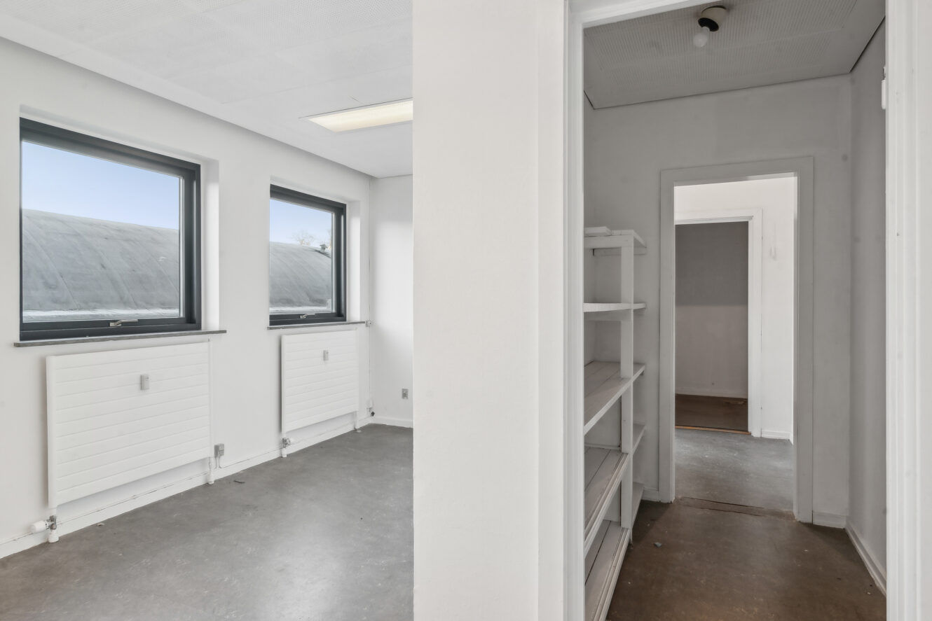 Et lille, tomt rum med to vinduer, hvide vægge og gråt gulv er synligt ved siden af en gang med indbyggede hvide hylder og flere rum.